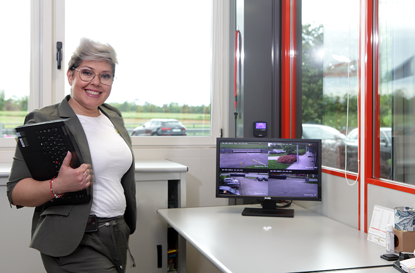EMCO Mitarbeiterin steht mit Laptop im Arm im Büro vor einem Bildschirm mit Überwachungsbildern